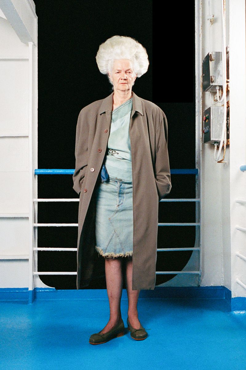 Mature adult female poses in a blue denim skirt and taupe coat on a cruise ship, with white railings and blue flooring visible.