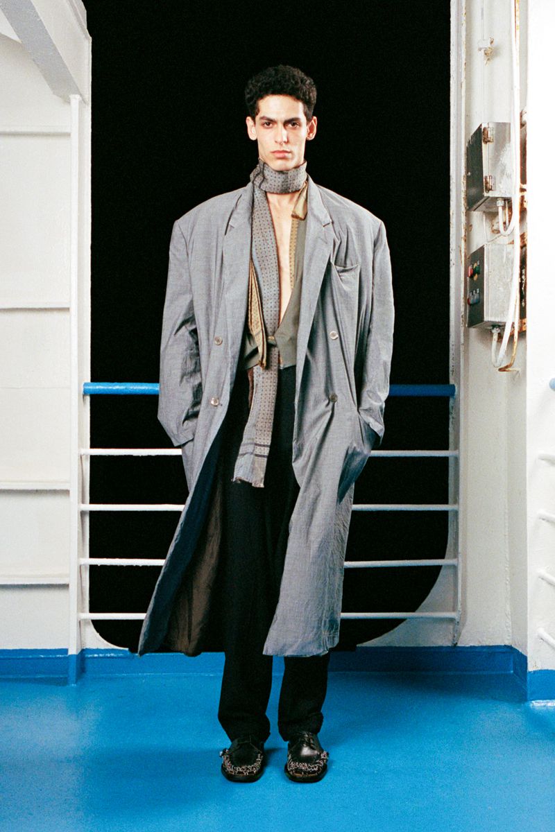 A stylish adult male in an oversized grey coat and black pants stands on a blue floor with a dark background in a fashion shot.