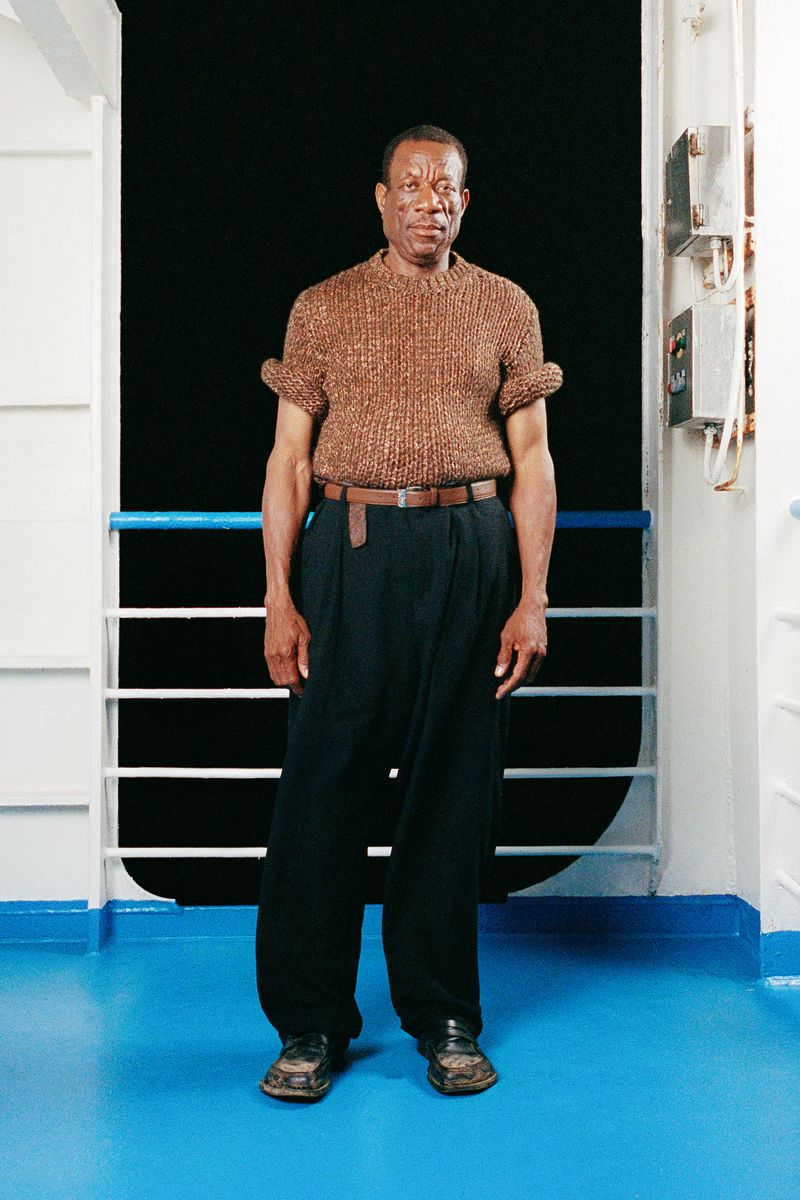 Full-length portrait of an adult Black man standing and posing on a ship with a brown knit shirt and navy pants, looking at the camera.