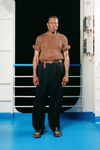 Full-length portrait of an adult Black man standing and posing on a ship with a brown knit shirt and navy pants, looking at the camera.
