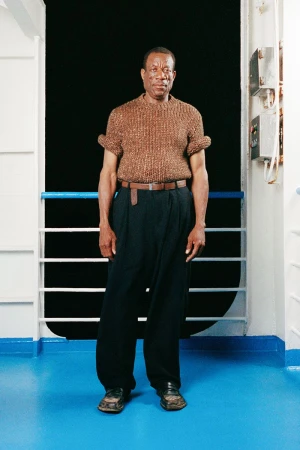 Full-length portrait of an adult Black man standing and posing on a ship with a brown knit shirt and navy pants, looking at the camera.
