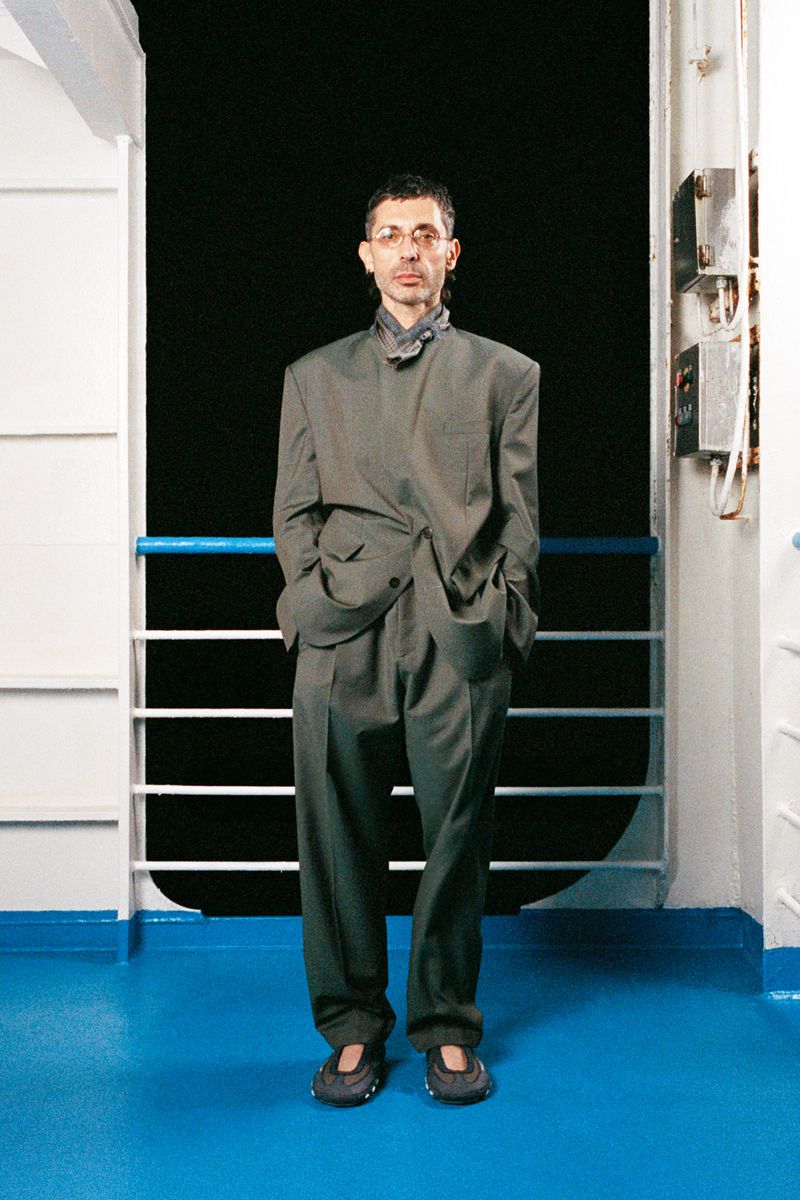 Adult man wearing oversized grey suit stands indoors against a black backdrop with blue railing. Formal wear fashion.
