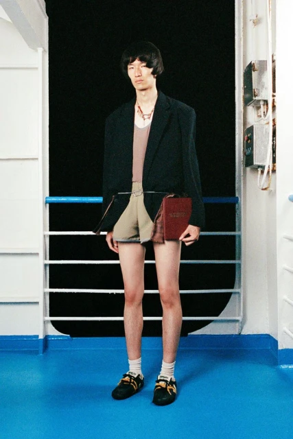 Young male fashion model poses on ship deck in blazer and shorts, holding a book, featuring blue deck and black background.