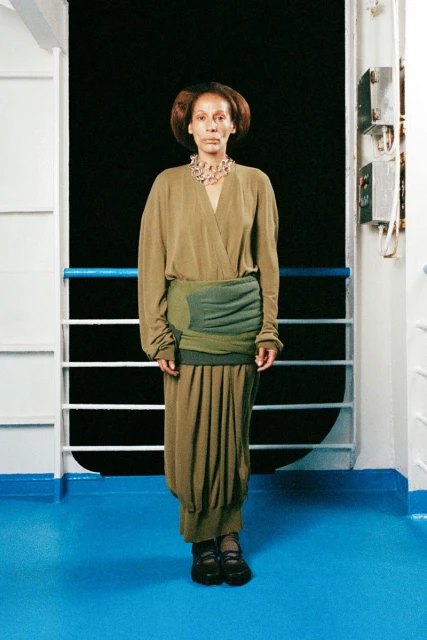 A full shot of an adult female with brown hair, wearing a beige wrap dress and large necklace, standing on a blue floor with a white background.