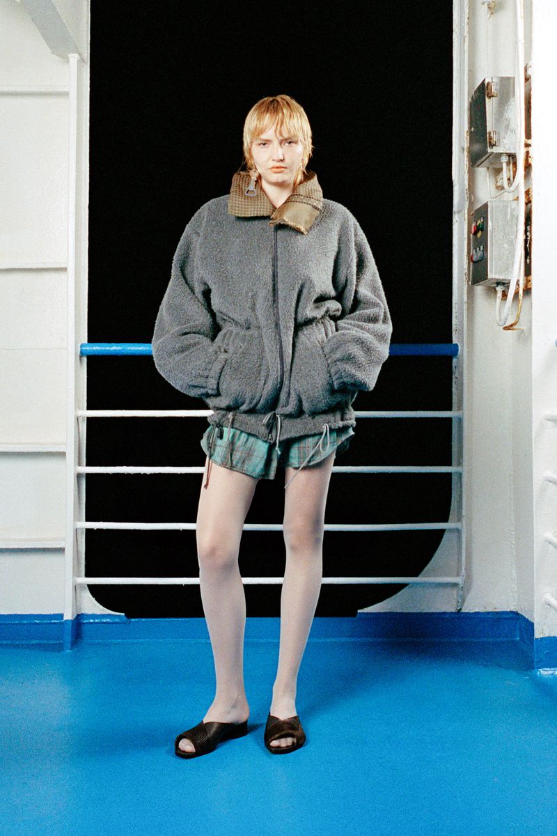 A young woman with blonde hair posing in a grey jacket, shorts and slides against a boat railing and black background, medium shot.