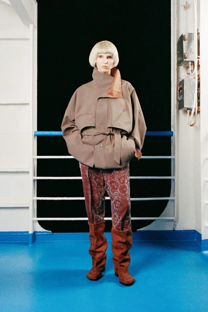 Fashion model in a brown jacket, patterned pants, and boots poses on a ship with a dark background, creating an editorial style image.