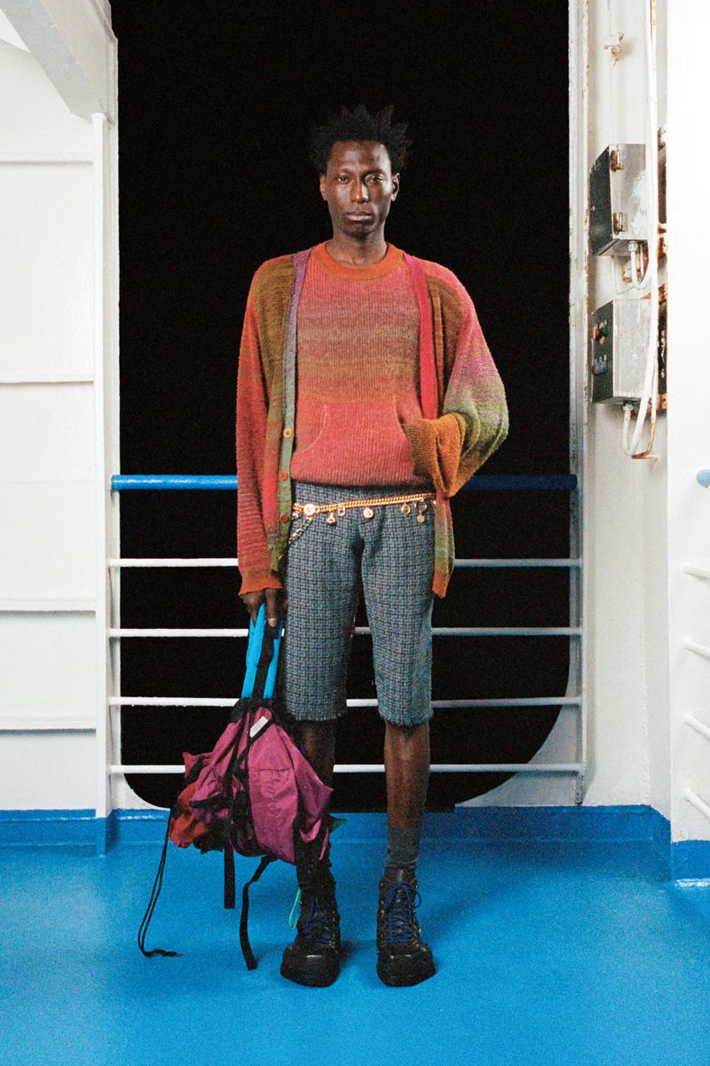 Full shot of a Black male model wearing casual fashion standing on a ferry boat with a backpack in hand, looking serious and stylish.