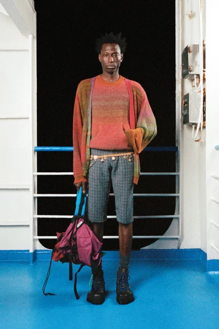 Full shot of a Black male model wearing casual fashion standing on a ferry boat with a backpack in hand, looking serious and stylish.