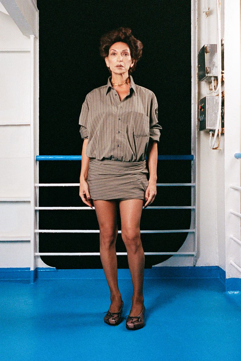 Full shot of an adult woman in an unbuttoned striped shirt-dress and brown high heels, posing on a blue and white boat deck in a studio.