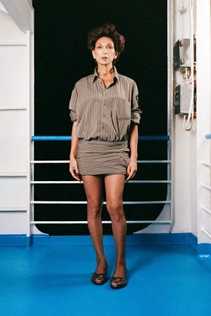 Full shot of an adult woman in an unbuttoned striped shirt-dress and brown high heels, posing on a blue and white boat deck in a studio.