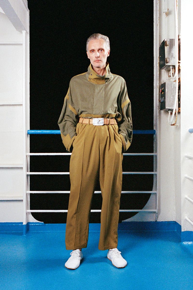 Adult male in stylish khaki and olive outfit with white shoes, posing on a ship deck with a black background, creating a fashion editorial.