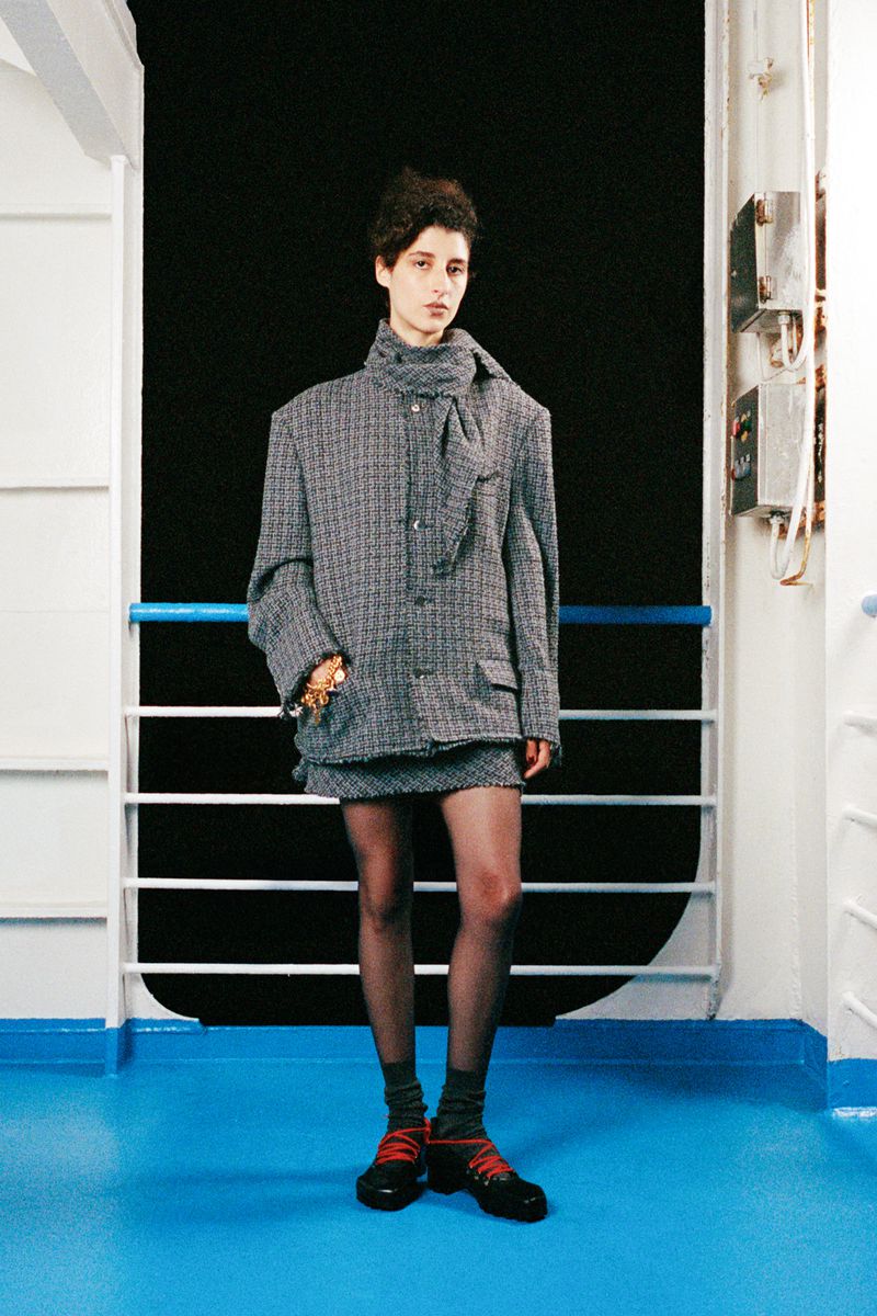 Fashion shot of a model in a tweed jacket and skirt, posing indoors against a white wall with blue trim and a black backdrop.