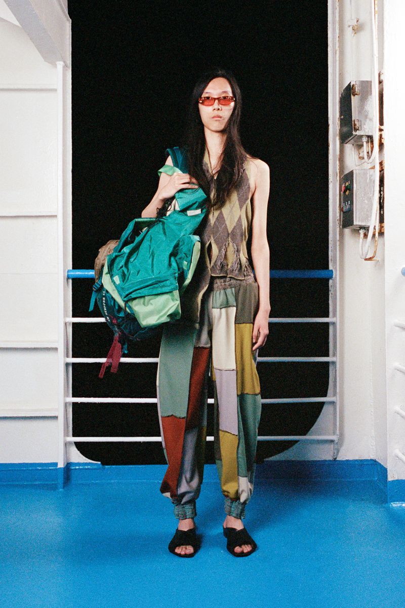 Fashionable adult with backpack poses in patchwork pants and orange glasses against a white railing and dark background, full shot.