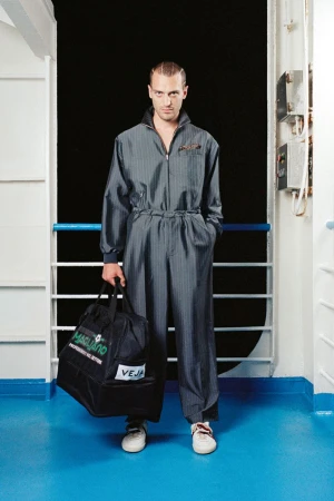 Model wearing a grey zipped jumpsuit holding a black Magliano duffle bag against a ship railing and dark background.