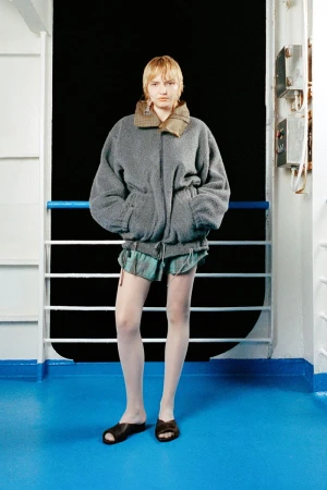 Model in an oversized grey fleece jacket, plaid shorts, and white tights posing on a blue ship deck against a dark background.