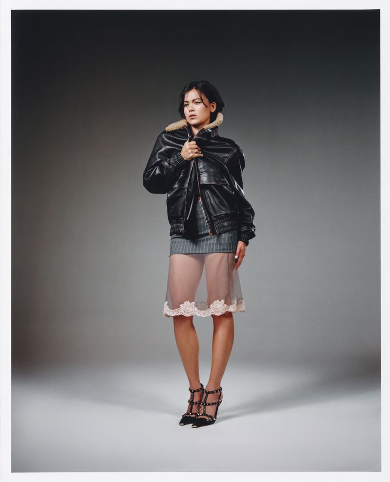 Full-length portrait of a woman wearing a black leather jacket and sheer skirt against a gradient grey studio backdrop.
