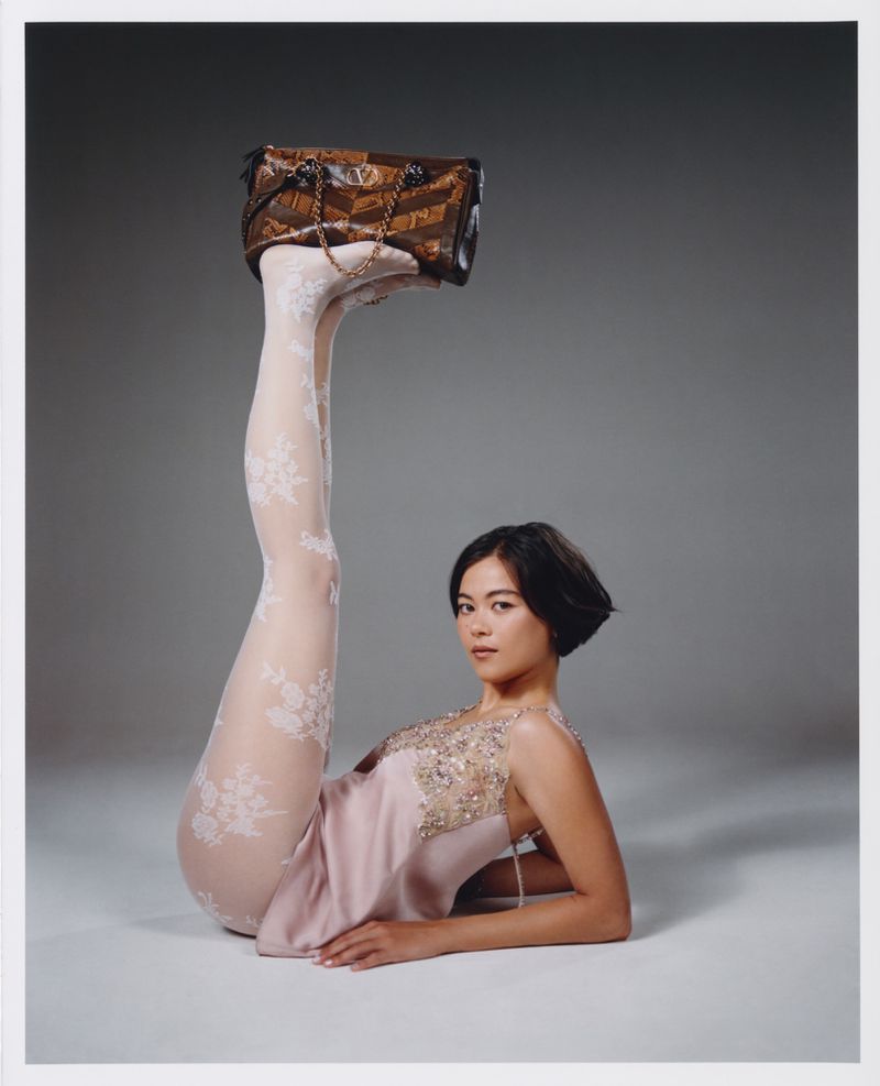 Lukita Maxwell reclining with legs raised in the air balancing a brown patterned handbag on her feet against a grey studio backdrop.