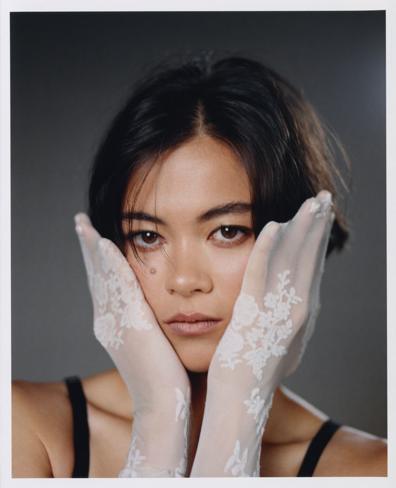 Lukita Maxwell posing with hands framing her face wearing sheer white floral lace gloves against a grey studio backdrop.