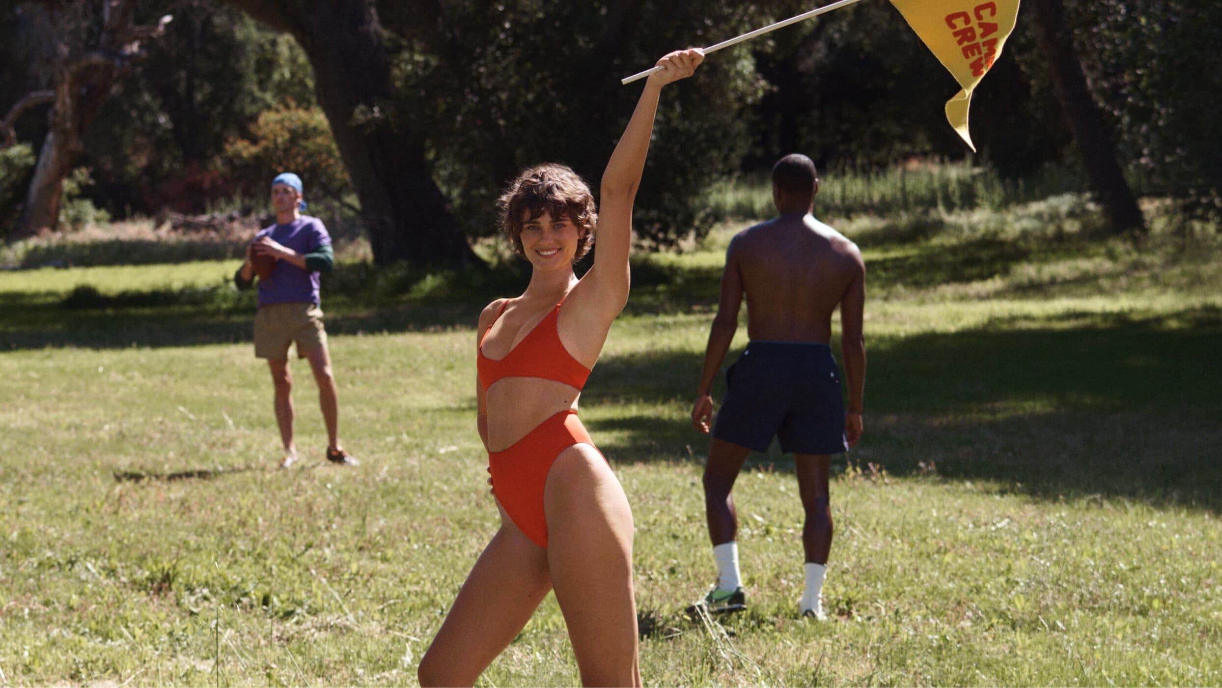 Woman in red bikini holding a yellow camp crew flag on a grassy field with other people standing in the background.
