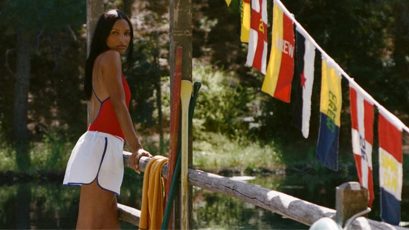 Model in a red swimsuit and white shorts leaning on a wooden dock with hanging flags