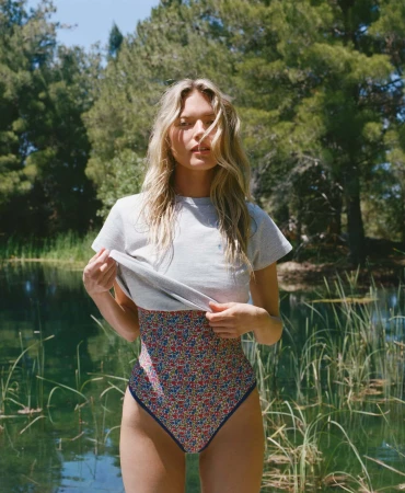 Martha Hunt lifting a grey t-shirt over a floral one-piece swimsuit while standing in front of a sunlit lake.