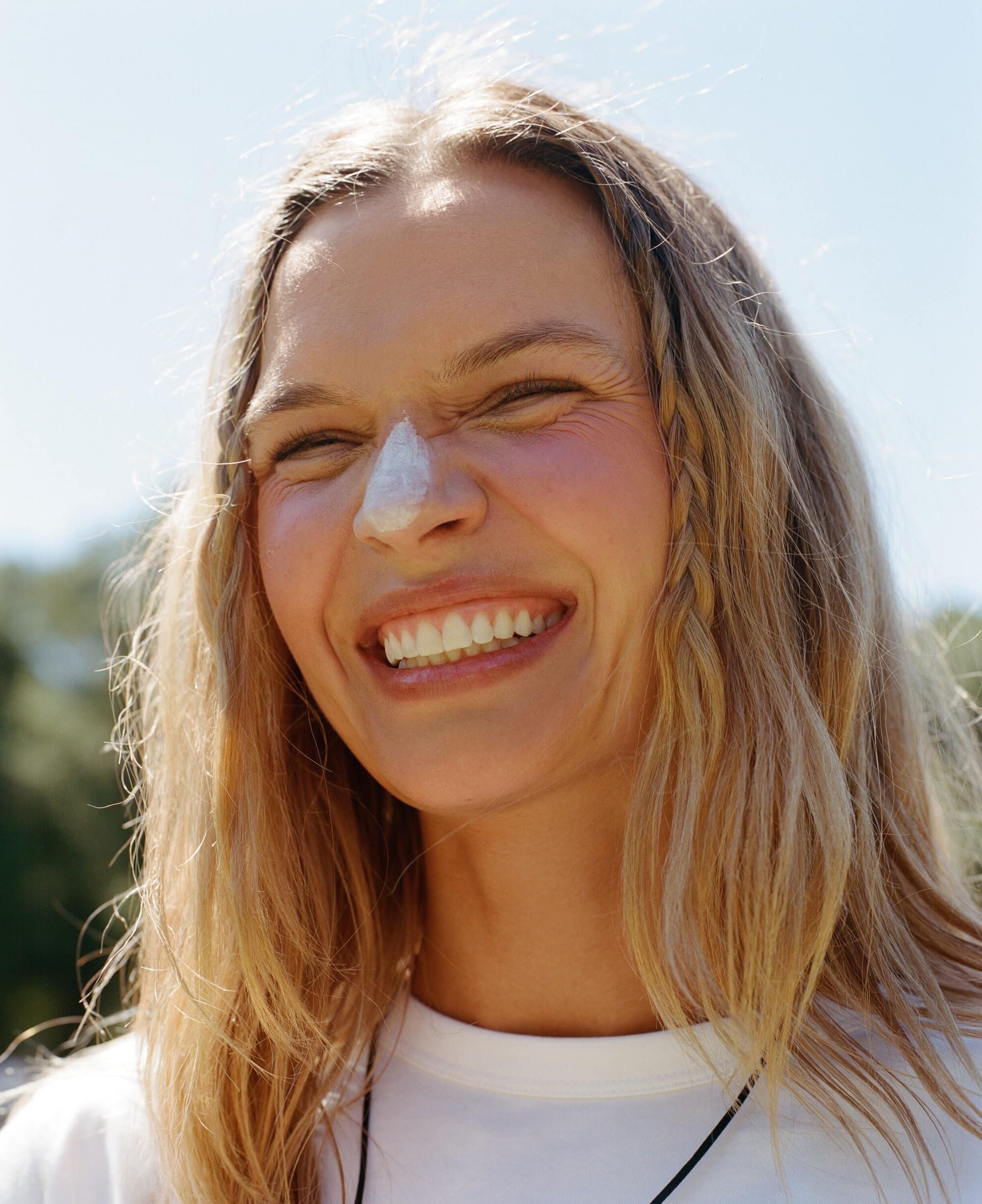 Josephine Skriver smiling with white zinc sunscreen on her nose in a white t-shirt under bright direct sunlight.