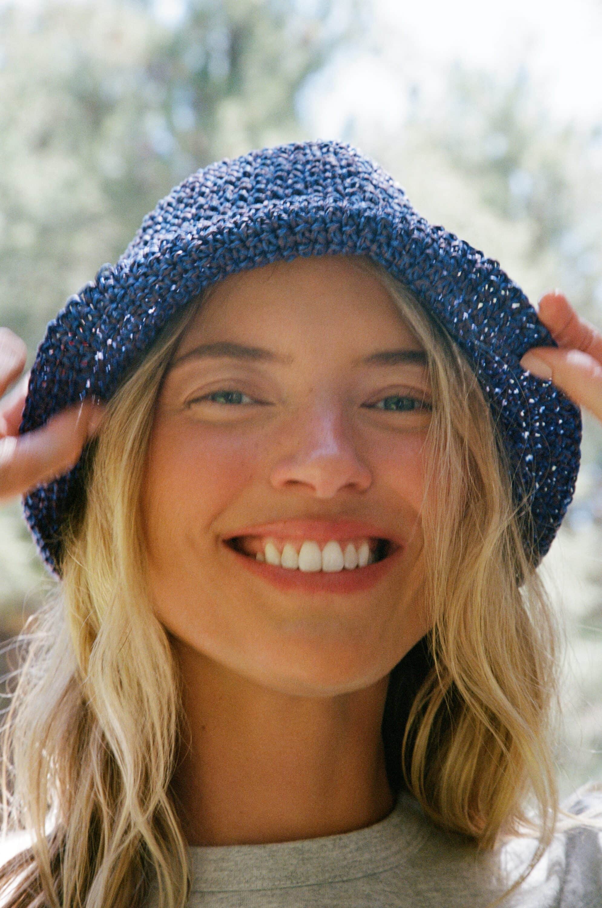 Close-up portrait of model Martha Hunt smiling while wearing a woven navy bucket hat outdoors in natural daylight.