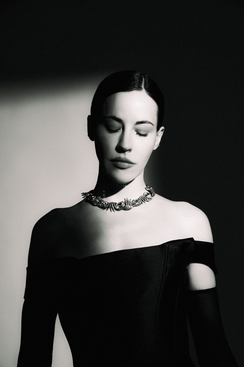 Anna Alimani wearing an off-the-shoulder black dress and ornate choker necklace in high-contrast black and white studio lighting.
