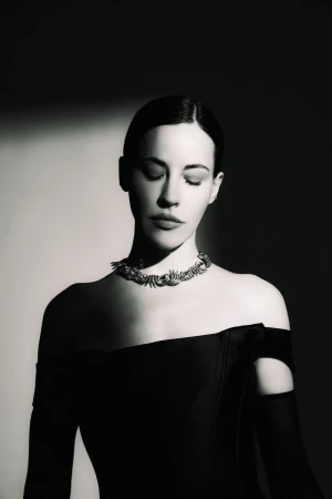 Anna Alimani wearing an off-the-shoulder black dress and ornate choker necklace in high-contrast black and white studio lighting.