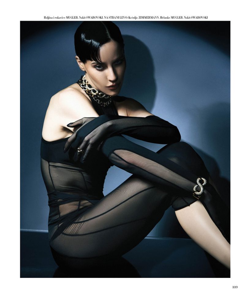 Model Anna Alimani seated in a sheer black Mugler bodysuit with long gloves and a statement necklace against a cool blue spotlight background.