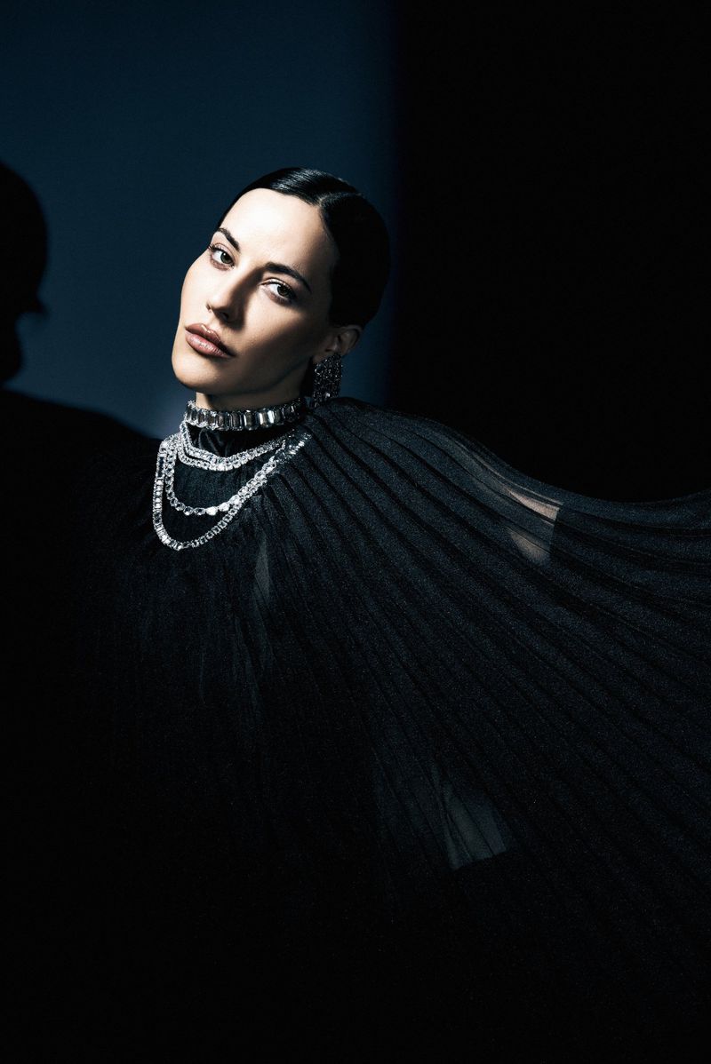 Anna Alimani wearing a pleated black cape and layered crystal choker necklace against a dark background.