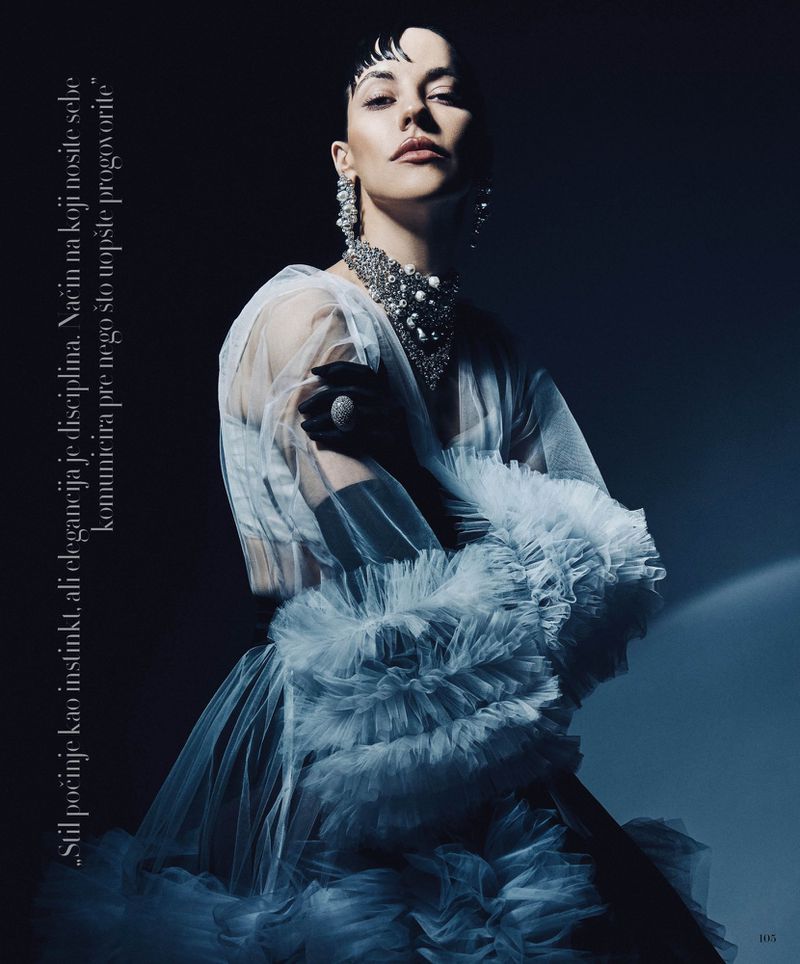 Model Anna Alimani poses in a ruffled tulle gown, jeweled choker, and black gloves against a dark background.