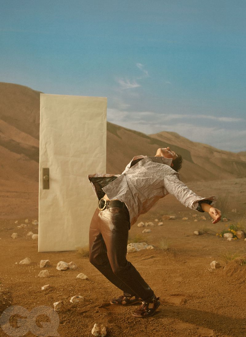 A man in a printed jacket and brown trousers leans dramatically backward in a desert next to a freestanding white door.