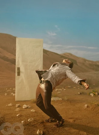 A man in a printed jacket and brown trousers leans dramatically backward in a desert next to a freestanding white door.