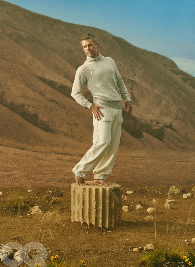 Actor Matt Damon standing barefoot on a classical stone column wearing a cream turtleneck sweater and sweatpants in an arid setting.