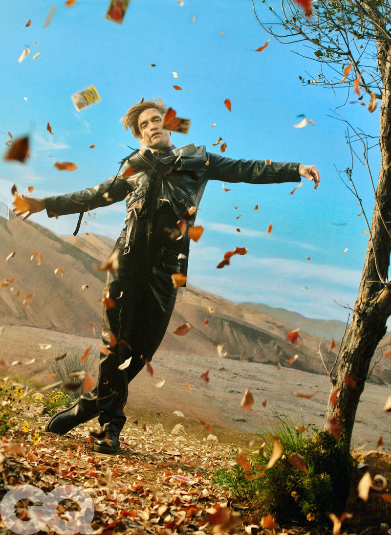 Robert Pattinson leaning back outdoors in a black leather jacket and pants surrounded by swirling autumn leaves and cards.