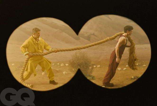 Matt Damon in yellow clothing and Tom Holland in brown pulling a thick rope seen through a binocular frame in an arid desert.