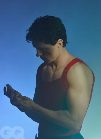 Tom Holland looking down at his hands wearing a red sleeveless top against a blue studio background.