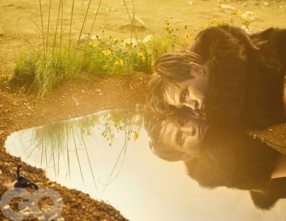 Actor Robert Pattinson in a dark fur coat lying on dry ground beside a puddle, looking at his water reflection.