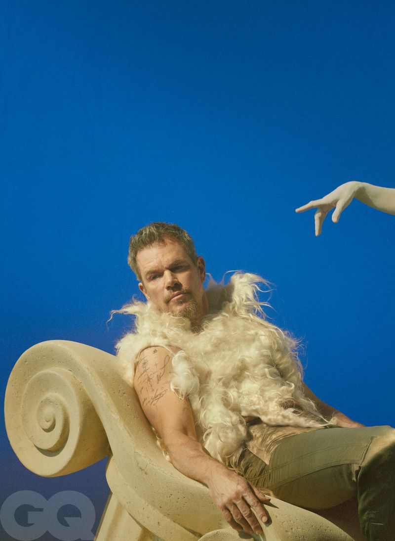 Matt Damon reclines on an ornate classical stone scroll wearing a fluffy cream vest against a vivid blue sky for GQ Magazine.