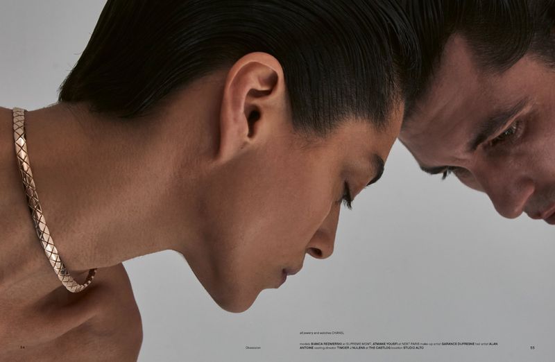 Close-up profile of a woman wearing a metallic woven necklace next to a man with slicked-back dark hair against a light grey background.