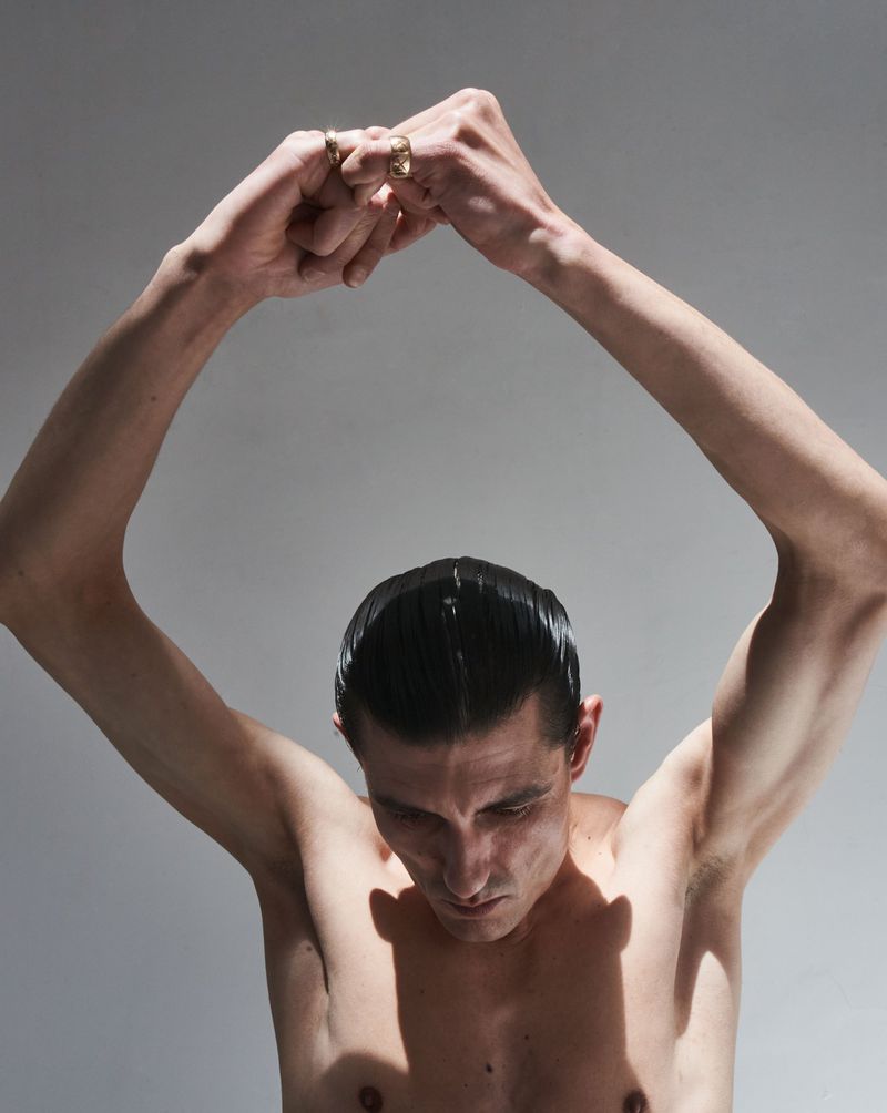Close-up portrait of a shirtless male model with hands clasped above his head against a neutral grey studio background