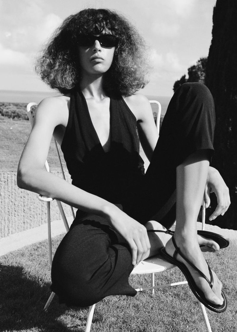 Model in sunglasses wearing a black halter top and trousers seated outdoors on a white wire chair in a black and white photograph