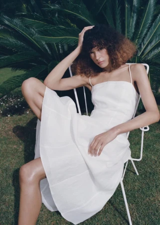 Model Prinnie Stott seated in a white wire chair wearing a white spaghetti-strap dress in front of lush green palm leaves.