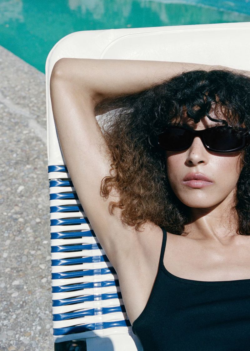Model Prinnie Stott wearing black sunglasses and a black swimsuit while reclining on a poolside lounge chair in bright sunlight.