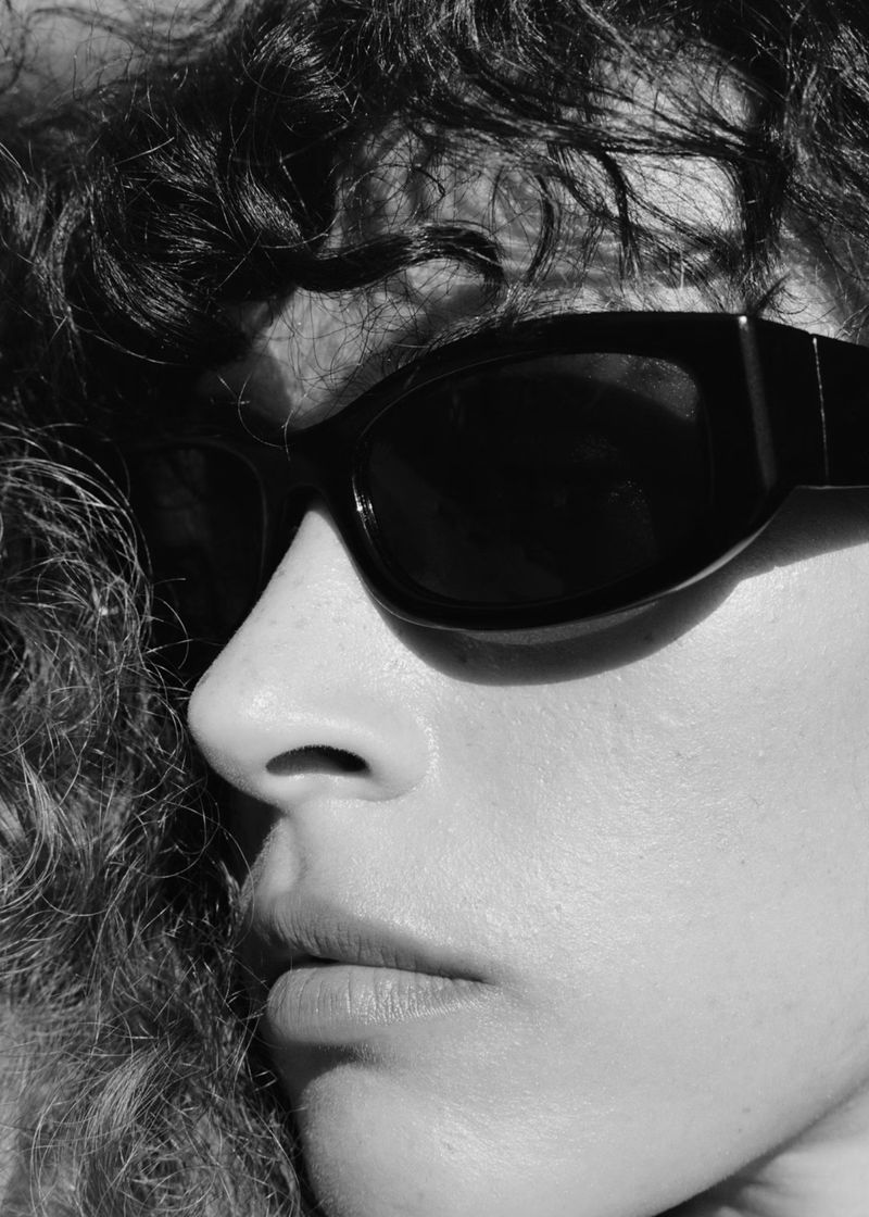 Black and white close-up portrait of a woman with curly hair wearing dark oval sunglasses under direct sunlight.