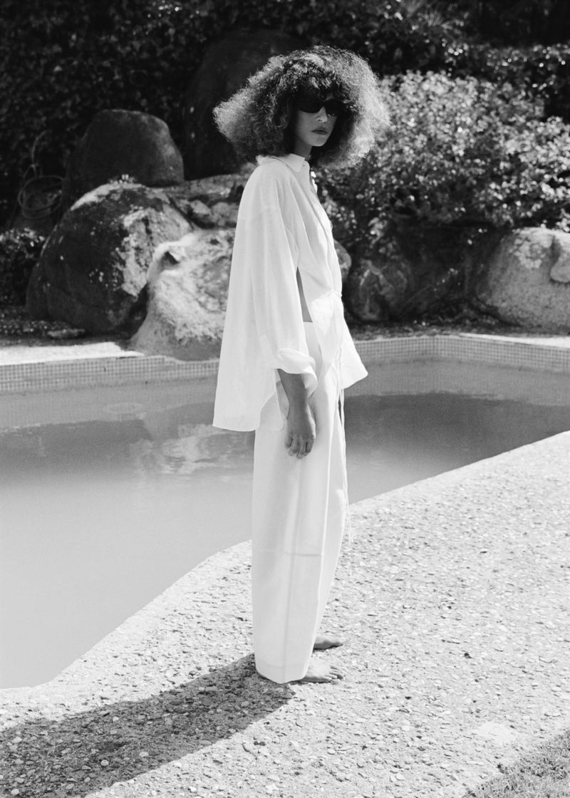 Model Prinnie Stott standing poolside in an oversized white shirt and wide-leg trousers wearing dark sunglasses.