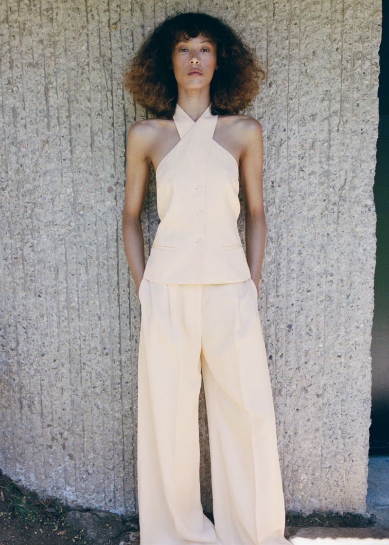 Model Prinnie Stott poses in a pale yellow halter top and matching wide-leg trousers against a textured concrete wall for Mango.