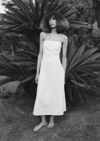 Model Prinnie Stott in a white spaghetti strap midi dress standing barefoot outdoors in front of lush palm fronds.