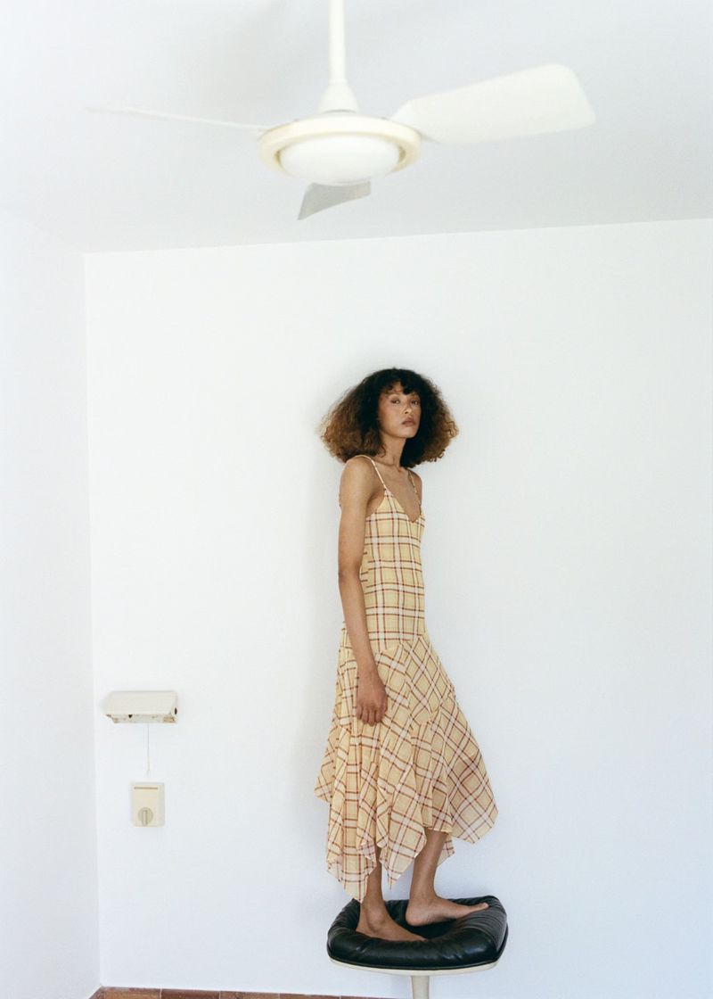 Model Prinnie Stott wearing a yellow plaid dress stands barefoot on a black stool beneath a white ceiling fan.
